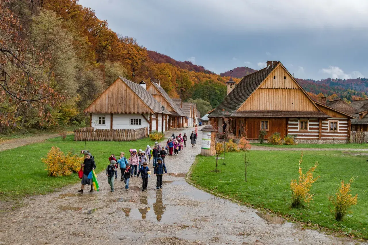 Dzieci zwiedzają skanseny w Polsce. Jesienne liście zdobią wzgórza za drewnianymi chatami.
